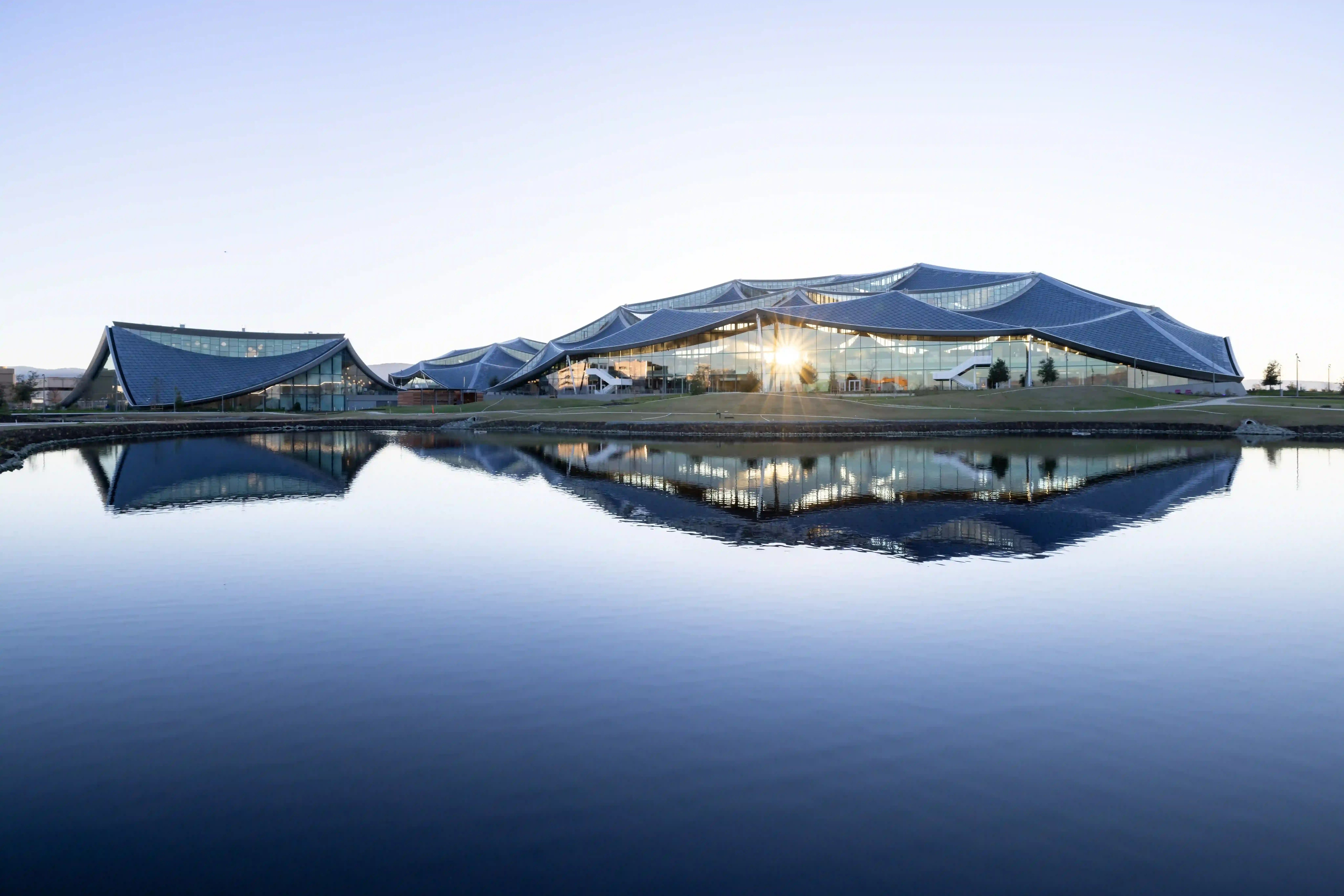 A view from afar of a large, tent-like glass building shining in the sun against a clear, pale blue sky.