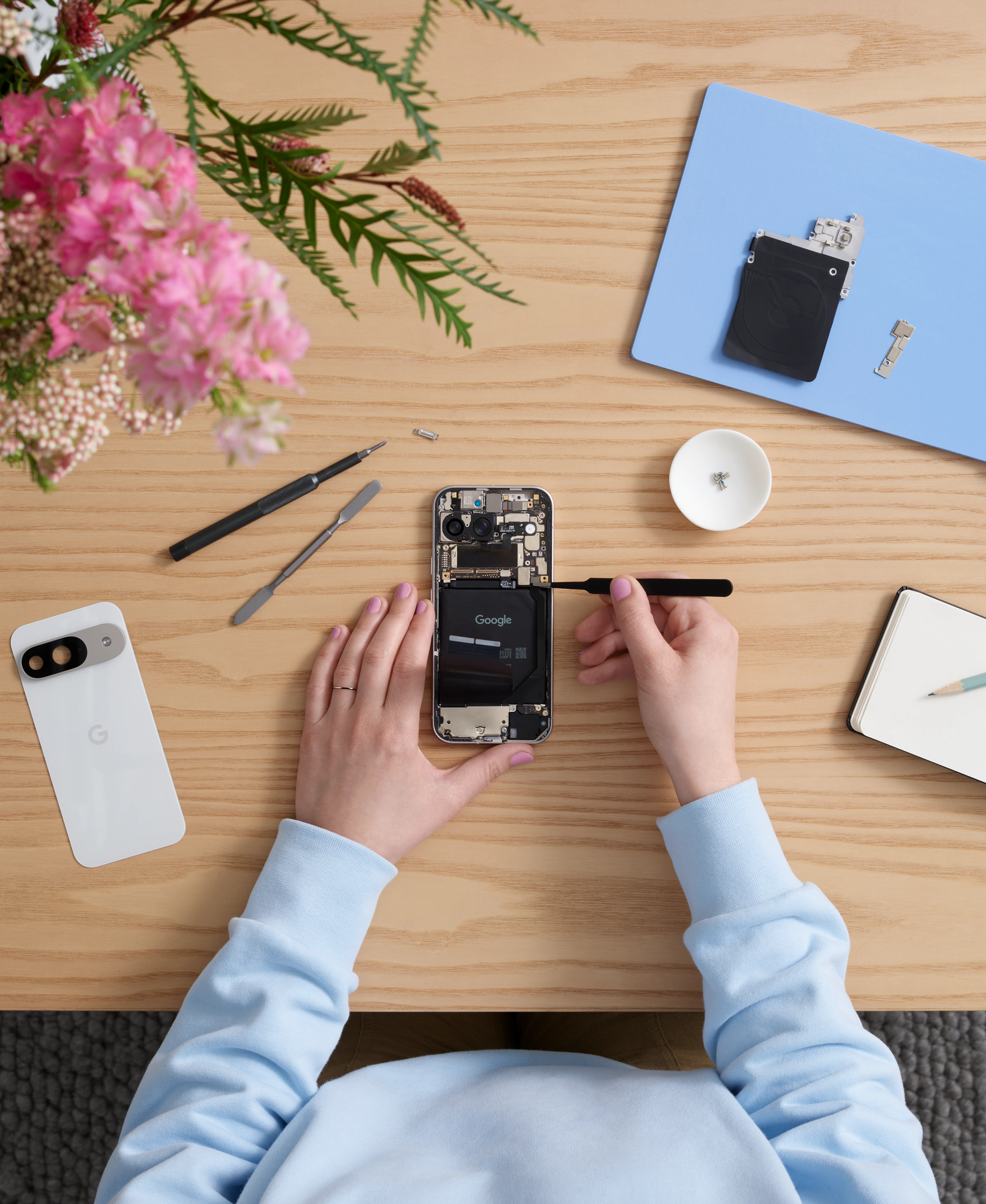 A person's hands are shown from a top-down perspective on a light wooden table, carefully disassembling a white smartphone with tools, alongside another white smartphone and pink flowers