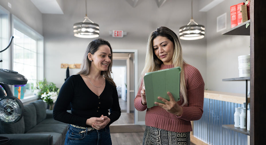 Two women are standing next to each other. The woman on the right is holding a tablet and discussing its content with the other woman. 