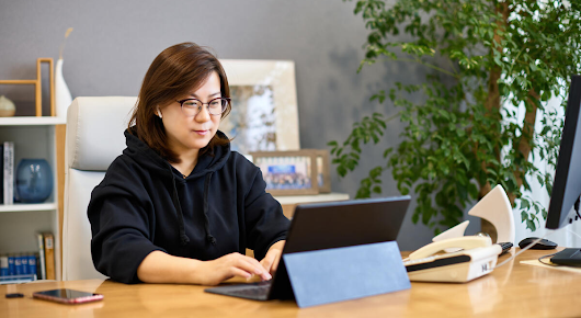 A woman wearing glasses and sitting at a desk is typing on a laptop. There is a mobile phone as well as a desktop phone on the table.