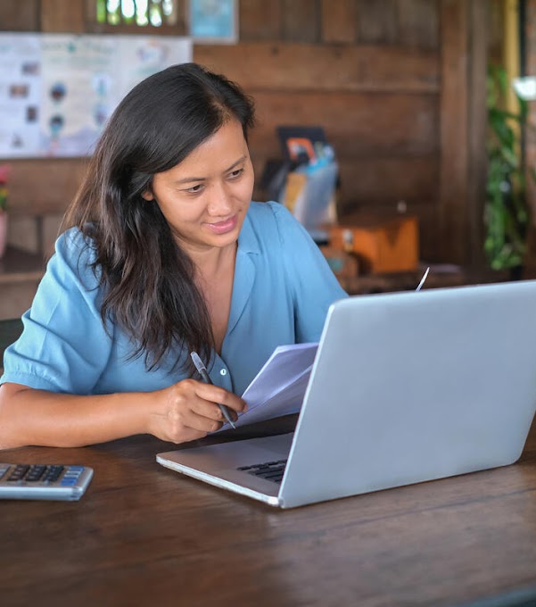 A woman is sitting at a table, holding papers and a pen. A laptop and a calculator can be found on the table in front of her.