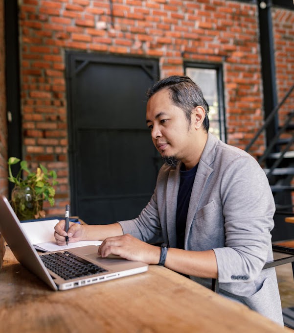 A man photographed from the side is sitting at a table, scrolling on his laptop with one hand, and taking notes in a notebook with the other.