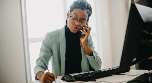 A woman with a green suit and glasses is sitting at a desk in front of a monitor. She is on the phone and writing in a notebook.