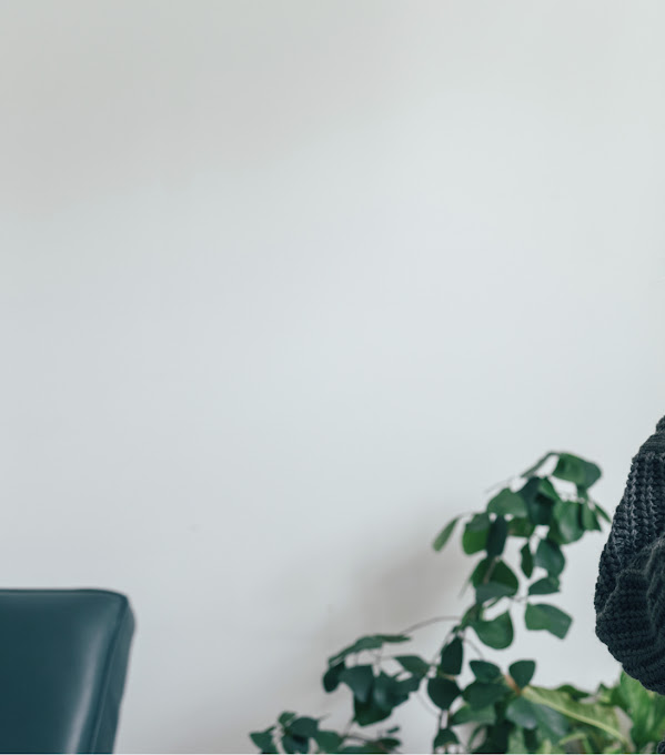 グレーのセーターを着た女性が手に持ったタブレットを見ている。女性の後ろには観葉植物、右手側には窓がある。