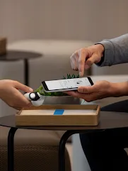 A hand holds Porcelain-colored Pixel Buds over a dark brown side table. Another hand points towards a phone screen.